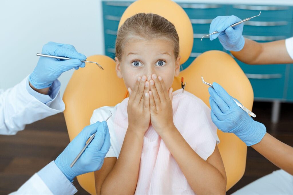 A nervous young girl with wide-open eyes covers her mouth while surrounded by dental tools. This image conveys common fears associated with dental visits and the need for compassionate, gentle pediatric dental care. 
