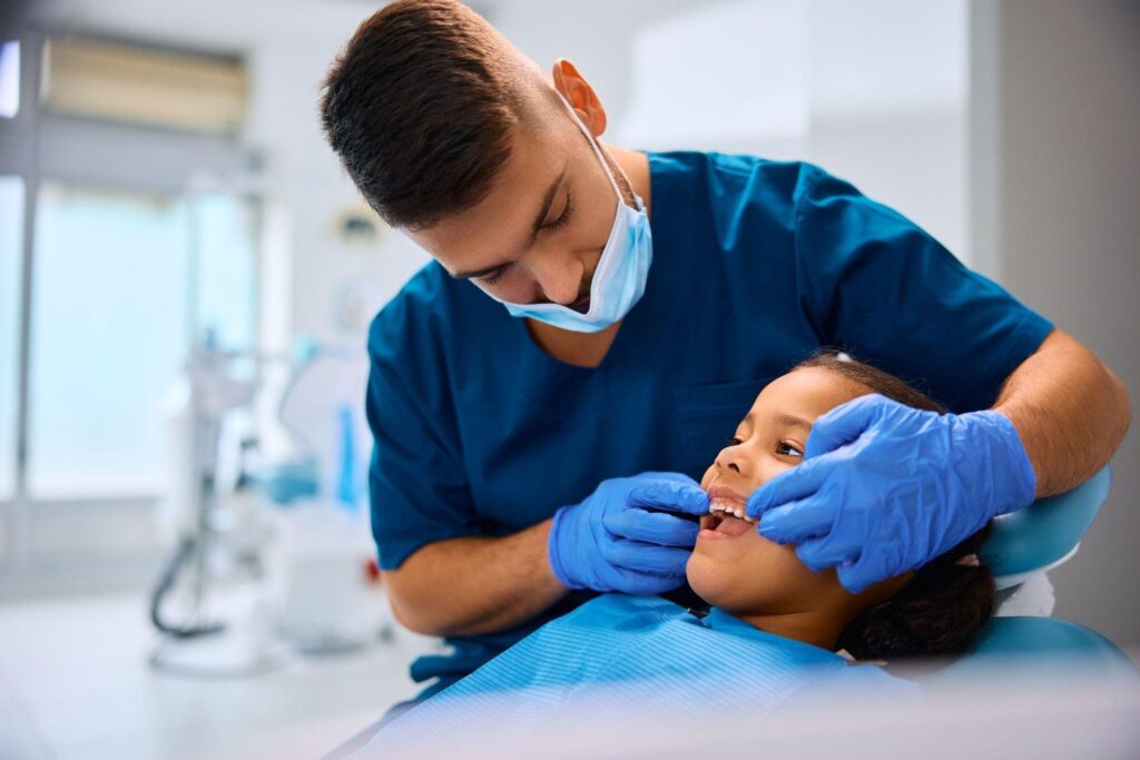 A young girl smiling while a pediatric dentist in blue scrubs examines her teeth. This image highlights a child-friendly dental environment and emphasizes the importance of routine pediatric dental checkups for maintaining oral health.