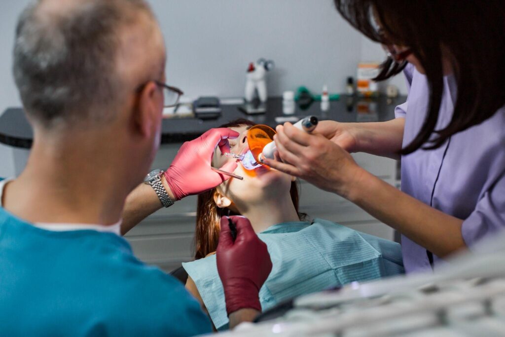 A dental team performs an in-office teeth whitening procedure on a young female patient. The dentist applies a whitening agent while using an LED or UV light to enhance the treatment. The patient wears orange protective glasses for eye safety. This professional whitening treatment provides fast and noticeable results.
